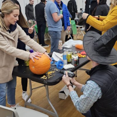 This image showcases a lively team building activity where participants are creating pumpkin racers. One person is working on carving or decorating a pumpkin, while another, wearing a festive witch's hat, appears to be using a power tool to attach components, possibly wheels, to the pumpkin. The setting has a group of people engaged in similar activities in the background, making it a fun and collaborative event, as part of a Halloween-themed competition. thumbnail
