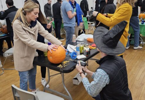 This image showcases a lively team building activity where participants are creating pumpkin racers. One person is working on carving or decorating a pumpkin, while another, wearing a festive witch's hat, appears to be using a power tool to attach components, possibly wheels, to the pumpkin. The setting has a group of people engaged in similar activities in the background, making it a fun and collaborative event, as part of a Halloween-themed competition.