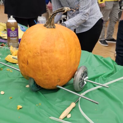 This image shows a pumpkin racer in the process of being assembled during a team building event. The pumpkin is fitted with metal rods serving as axles, and wheels are attached on either side. The scene captures the creativity of participants as they construct their racer, likely in preparation for a fun pumpkin race. The surrounding materials, drinks, and craft supplies add to the lively and collaborative atmosphere. thumbnail