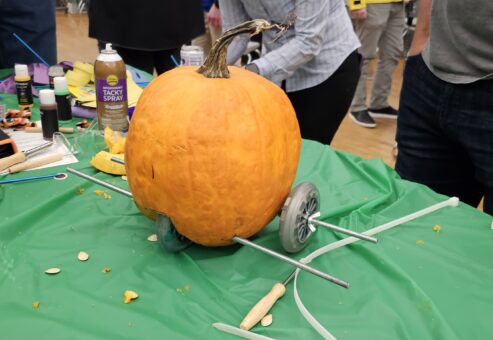 This image shows a pumpkin racer in the process of being assembled during a team building event. The pumpkin is fitted with metal rods serving as axles, and wheels are attached on either side. The scene captures the creativity of participants as they construct their racer, likely in preparation for a fun pumpkin race. The surrounding materials, drinks, and craft supplies add to the lively and collaborative atmosphere.