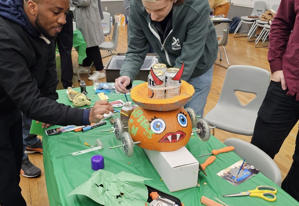 A group of individuals is working on a pumpkin racer, a fun Halloween-themed activity. The pumpkin racer has large, googly eyes and a wide mouth with pointed teeth painted on it. It is placed on wheels, ready for racing. The table is covered in tools and decorations such as scissors and crafting materials, while the team building participants focus on adding final touches to the pumpkin racer.
