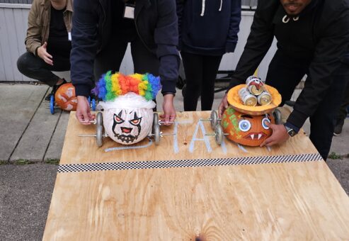 Two individuals are preparing pumpkin-themed cars for a race on a wooden surface. The pumpkin on the left is decorated with a colorful clown wig and a painted face, while the pumpkin on the right features a more monster-like design with sharp teeth and attached eyes. Both pumpkins have wheels attached, ready to race. A checkerboard line is visible on the board, marking the starting point of this Great Pumpkin Team Building Race.