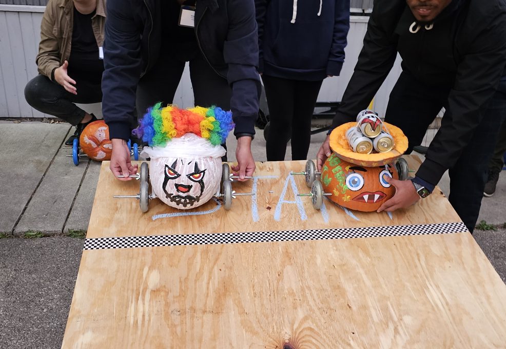 Two individuals are preparing pumpkin-themed cars for a race on a wooden surface. The pumpkin on the left is decorated with a colorful clown wig and a painted face, while the pumpkin on the right features a more monster-like design with sharp teeth and attached eyes. Both pumpkins have wheels attached, ready to race. A checkerboard line is visible on the board, marking the starting point of this Great Pumpkin Team Building Race.