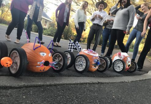 This image captures a fun moment from a pumpkin team building race event. Three creatively decorated pumpkins are lined up, each mounted on wheels to form pumpkin racers. The racers have unique designs: one appears to have been styled like a cat, with pipe cleaner whiskers and ears. Others are adorned with checkered flags and various festive decorations. In the background, participants are smiling and enjoying the activity, clearly excited about the upcoming race.