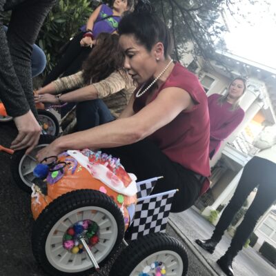 A participant is in the middle of a pumpkin race team building event. The pumpkin racer is decorated with colorful pom-poms, pipe cleaners, and a miniature checkered flag, symbolizing the competitive yet festive atmosphere of the race. The person is focused on adjusting the wheels of the creatively designed pumpkin racer, which is fitted with large wheels for mobility. thumbnail