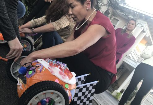 A participant is in the middle of a pumpkin race team building event. The pumpkin racer is decorated with colorful pom-poms, pipe cleaners, and a miniature checkered flag, symbolizing the competitive yet festive atmosphere of the race. The person is focused on adjusting the wheels of the creatively designed pumpkin racer, which is fitted with large wheels for mobility.