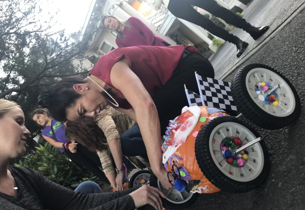 A participant is in the middle of a pumpkin race team building event. The pumpkin racer is decorated with colorful pom-poms, pipe cleaners, and a miniature checkered flag, symbolizing the competitive yet festive atmosphere of the race. The person is focused on adjusting the wheels of the creatively designed pumpkin racer, which is fitted with large wheels for mobility.