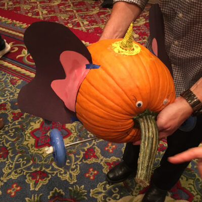 This image features a whimsical pumpkin racer designed to resemble an elephant. The pumpkin is decorated with large, pink and black ears attached to the sides and has a small horn or crown on top, adding a fun detail. The pumpkin’s stem is positioned to mimic an elephant's trunk, and googly eyes have been added for an expressive touch. The racer is equipped with wheels, ready for the competition. The colorful, patterned carpet background adds to the playful atmosphere of this creative team building event. thumbnail