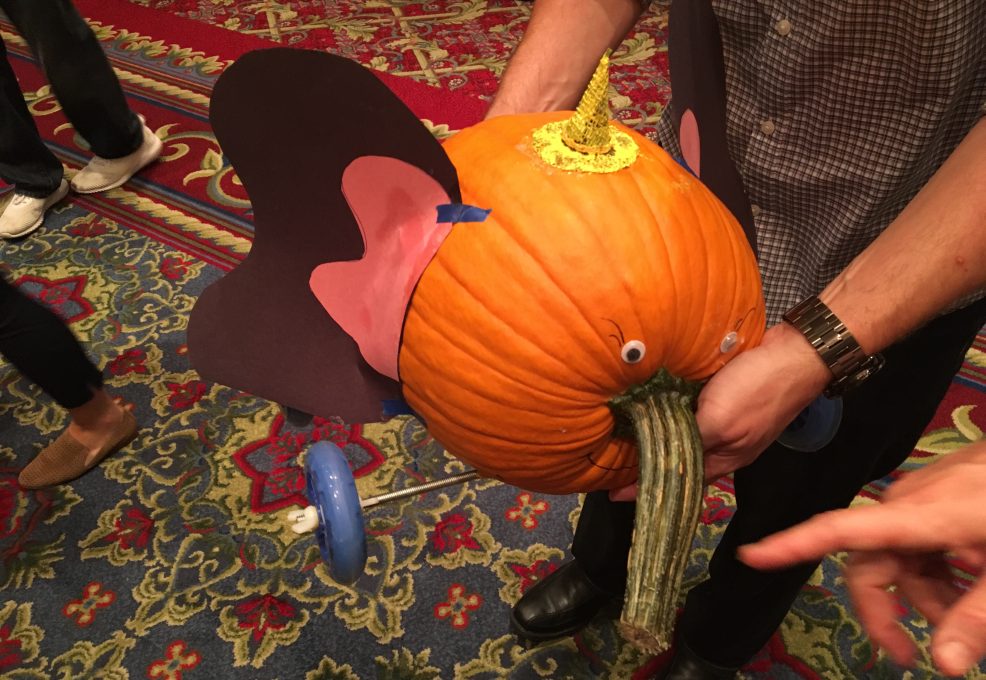 This image features a whimsical pumpkin racer designed to resemble an elephant. The pumpkin is decorated with large, pink and black ears attached to the sides and has a small horn or crown on top, adding a fun detail. The pumpkin’s stem is positioned to mimic an elephant's trunk, and googly eyes have been added for an expressive touch. The racer is equipped with wheels, ready for the competition. The colorful, patterned carpet background adds to the playful atmosphere of this creative team building event.