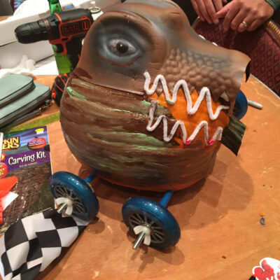 This image showcases a creatively designed pumpkin racer. The pumpkin has been intricately transformed to resemble a fierce creature, possibly inspired by a dinosaur or dragon, with painted scales and a set of sharp teeth. The design is very detailed, with wheels attached to the bottom for racing. A power drill and a pumpkin carving kit are visible in the background, suggesting that the team has been hard at work on this impressive creation. thumbnail