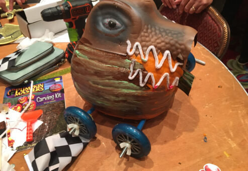 This image showcases a creatively designed pumpkin racer. The pumpkin has been intricately transformed to resemble a fierce creature, possibly inspired by a dinosaur or dragon, with painted scales and a set of sharp teeth. The design is very detailed, with wheels attached to the bottom for racing. A power drill and a pumpkin carving kit are visible in the background, suggesting that the team has been hard at work on this impressive creation.