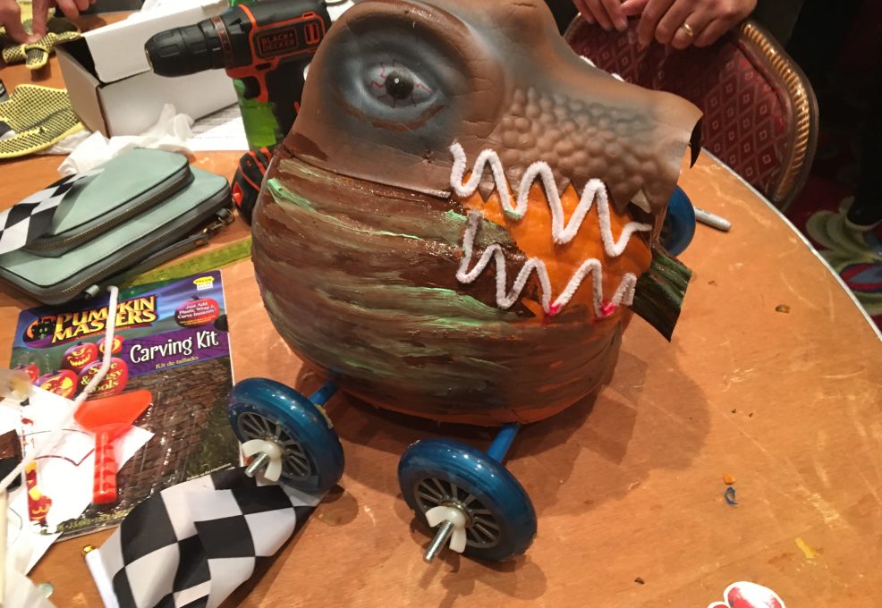This image showcases a creatively designed pumpkin racer. The pumpkin has been intricately transformed to resemble a fierce creature, possibly inspired by a dinosaur or dragon, with painted scales and a set of sharp teeth. The design is very detailed, with wheels attached to the bottom for racing. A power drill and a pumpkin carving kit are visible in the background, suggesting that the team has been hard at work on this impressive creation.