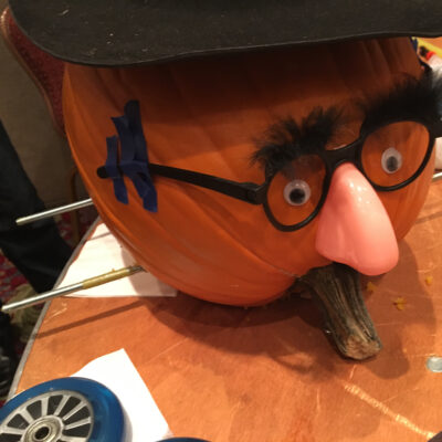 This image shows a creatively decorated pumpkin racer featuring a humorous face. The pumpkin has been outfitted with a comical disguise: a fake nose, glasses, and bushy eyebrows, giving it a playful personality. Axles for the wheels are visible, suggesting that this pumpkin is ready to be part of the pumpkin race. The setup adds a whimsical element to the team building activity, blending creativity with competition. thumbnail