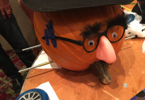 This image shows a creatively decorated pumpkin racer featuring a humorous face. The pumpkin has been outfitted with a comical disguise: a fake nose, glasses, and bushy eyebrows, giving it a playful personality. Axles for the wheels are visible, suggesting that this pumpkin is ready to be part of the pumpkin race. The setup adds a whimsical element to the team building activity, blending creativity with competition.