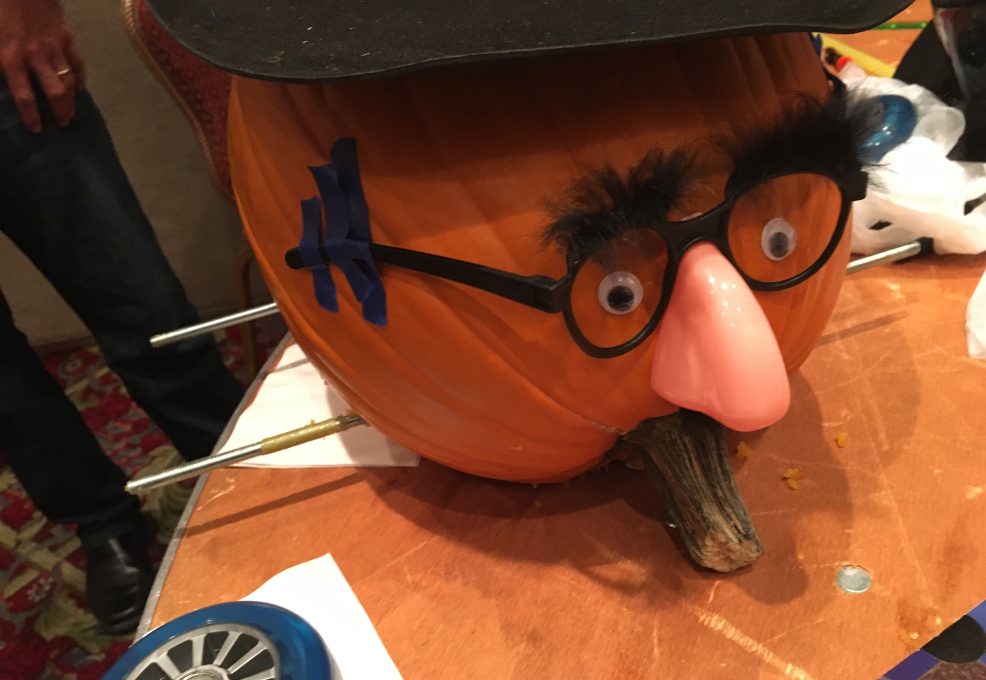 This image shows a creatively decorated pumpkin racer featuring a humorous face. The pumpkin has been outfitted with a comical disguise: a fake nose, glasses, and bushy eyebrows, giving it a playful personality. Axles for the wheels are visible, suggesting that this pumpkin is ready to be part of the pumpkin race. The setup adds a whimsical element to the team building activity, blending creativity with competition.
