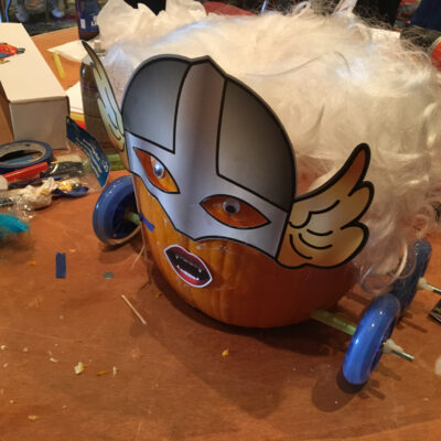 This image showcases a creatively designed pumpkin racer. The pumpkin has been transformed with a Viking-style mask, featuring wings on the side and dramatic facial expressions. The pumpkin is adorned with a fluffy white wig, adding a playful element to the design. The racer is fitted with blue wheels, and the tools and materials scattered around the workbench suggest that the team is in the midst of finalizing their creation. This fun and imaginative design emphasizes the lighthearted competition of the pumpkin race team building event. thumbnail