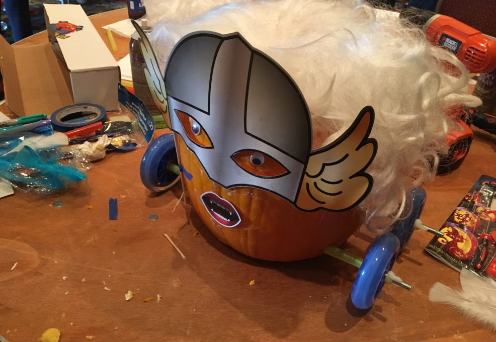 This image showcases a creatively designed pumpkin racer. The pumpkin has been transformed with a Viking-style mask, featuring wings on the side and dramatic facial expressions. The pumpkin is adorned with a fluffy white wig, adding a playful element to the design. The racer is fitted with blue wheels, and the tools and materials scattered around the workbench suggest that the team is in the midst of finalizing their creation. This fun and imaginative design emphasizes the lighthearted competition of the pumpkin race team building event.