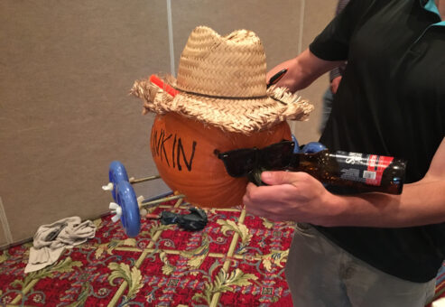 This image shows a creatively designed pumpkin racer, styled with personality. The pumpkin is adorned with a straw hat and sunglasses, giving it a cool, laid-back look. One of the wheels appears to be part of a toy or plastic assembly, attached to make the pumpkin mobile. The pumpkin also seems to be holding a bottle, adding to the humorous, festive vibe. It’s a fun and engaging way to bring creativity and teamwork to a pumpkin racing team building event.