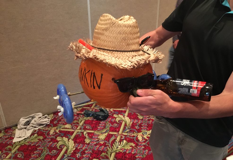 This image shows a creatively designed pumpkin racer, styled with personality. The pumpkin is adorned with a straw hat and sunglasses, giving it a cool, laid-back look. One of the wheels appears to be part of a toy or plastic assembly, attached to make the pumpkin mobile. The pumpkin also seems to be holding a bottle, adding to the humorous, festive vibe. It’s a fun and engaging way to bring creativity and teamwork to a pumpkin racing team building event.