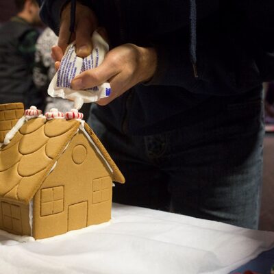 Close-up of a person decorating a gingerbread house by piping white icing on the roof and adding peppermint candies, during a holiday-themed team building event. thumbnail