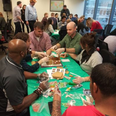 A team of six participants gathered around a table covered with a green cloth, actively building a gingerbread house during a team building competition. The group is engaged in the process, wearing gloves, and using various candy and gingerbread materials to assemble their house. Other teams can be seen in the background participating in the same activity in a lively and collaborative setting. thumbnail