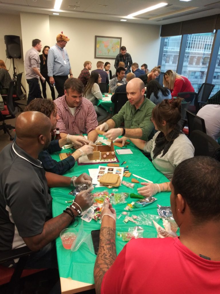 A team of six participants gathered around a table covered with a green cloth, actively building a gingerbread house during a team building competition. The group is engaged in the process, wearing gloves, and using various candy and gingerbread materials to assemble their house. Other teams can be seen in the background participating in the same activity in a lively and collaborative setting.