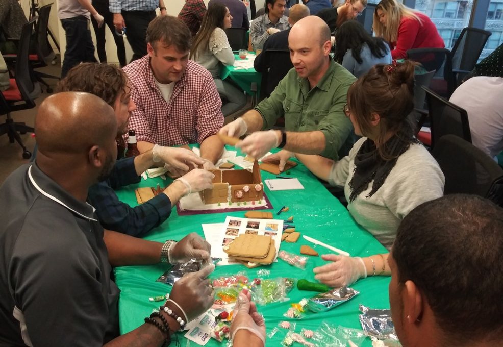 A team of six participants gathered around a table covered with a green cloth, actively building a gingerbread house during a team building competition. The group is engaged in the process, wearing gloves, and using various candy and gingerbread materials to assemble their house. Other teams can be seen in the background participating in the same activity in a lively and collaborative setting.