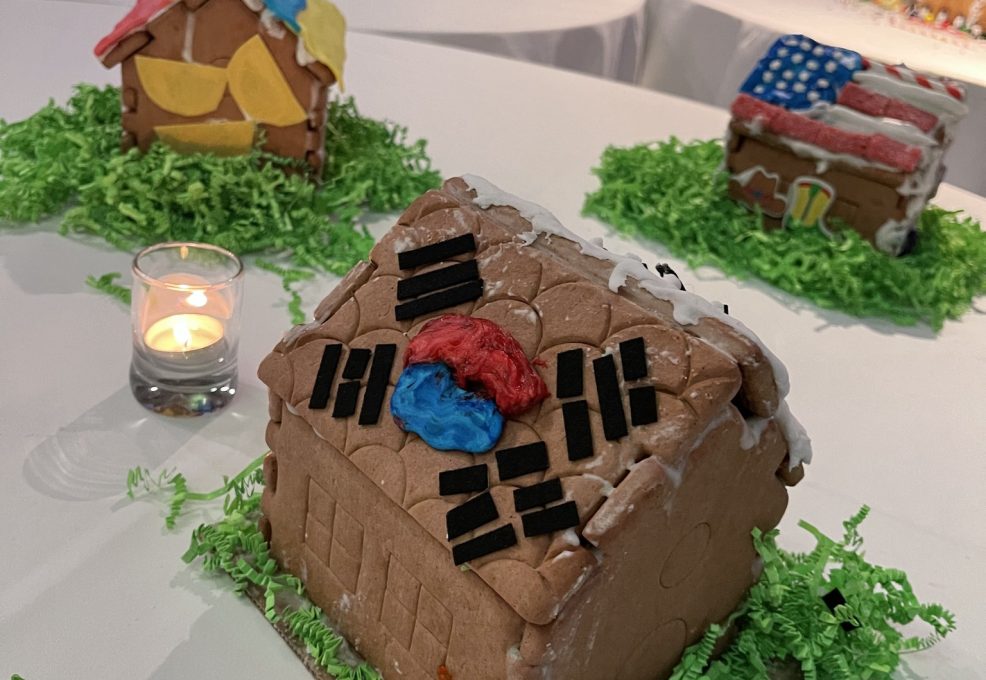 A gingerbread house decorated with icing and black licorice pieces, featuring the South Korean flag symbol in the center of the roof. The house is placed on a white table with green decorative grass around it, alongside a small lit candle. Other uniquely decorated gingerbread houses are visible in the background as part of a team building event.