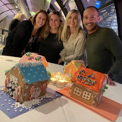 A group of four participants smiling and standing behind a table displaying three colorfully decorated gingerbread houses during a gingerbread house team building competition. The table is decorated with small lit candles, adding a cozy touch to the festive atmosphere.