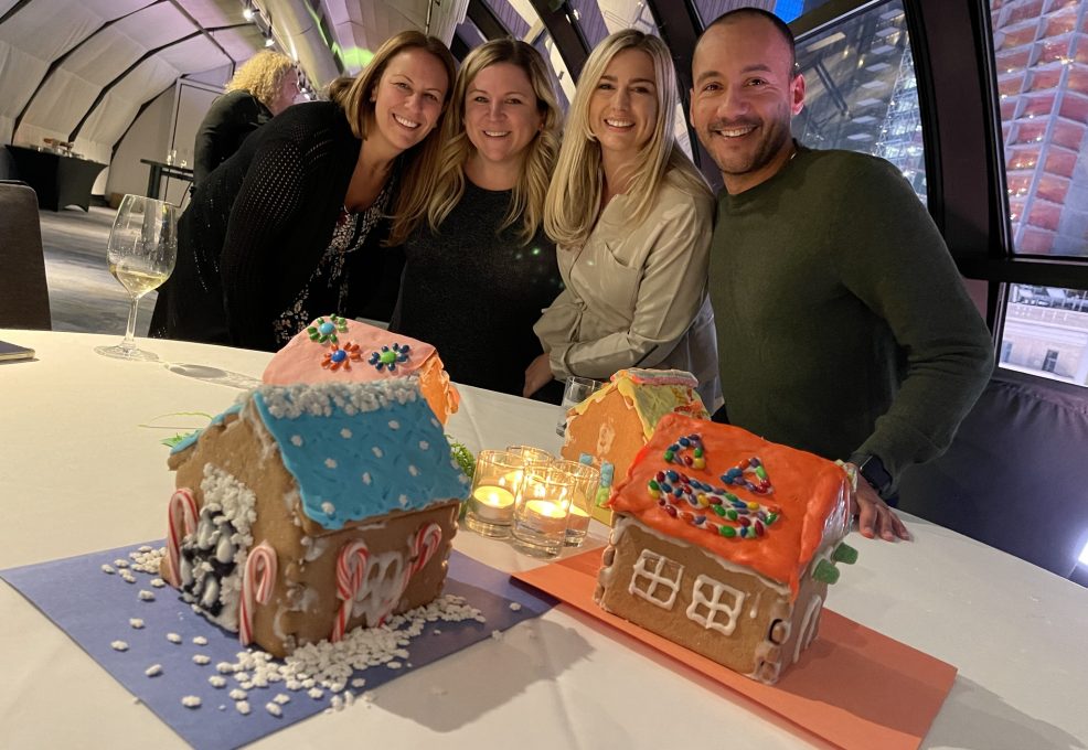 A group of four participants smiling and standing behind a table displaying three colorfully decorated gingerbread houses during a gingerbread house team building competition. The table is decorated with small lit candles, adding a cozy touch to the festive atmosphere.