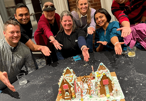 A group of seven enthusiastic participants proudly presenting their gingerbread house creation during a team building event. The gingerbread structure is decorated with icing, candy canes, and colorful candies. The team members are smiling and extending their hands toward the gingerbread house.