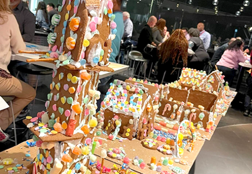 A display of intricately decorated gingerbread houses lined up during a team building competition. The houses are adorned with colorful candies, icing, and sprinkles, with one particularly tall gingerbread structure standing out in the foreground.