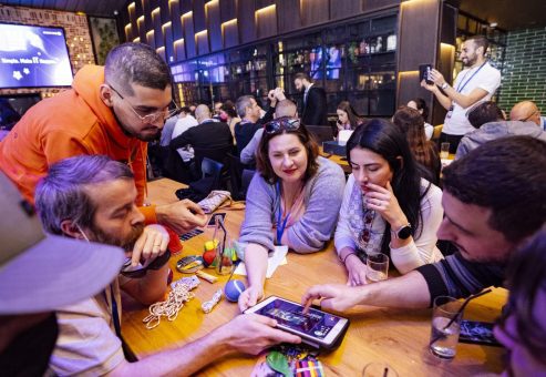 Team members huddle around their iPad, strategizing and collaborating to solve various puzzles, surrounded by game materials like hacky sacks and play dough. The fast-paced, interactive environment fuels teamwork and creative thinking.