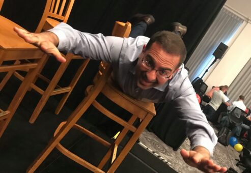 A participant fully embraces the fun and physical aspect of the game, executing a dramatic chair stunt while surrounded by a team engaged in the lively atmosphere. This highlights the playful, energetic spirit of the event.