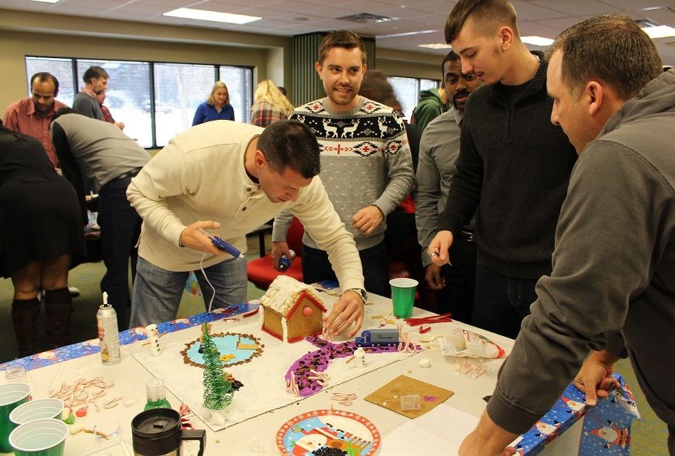 A group of men working together on decorating a gingerbread house during a team building event. One man uses a hot glue gun to assemble pieces, while others discuss and add decorations to the house on a festive, holiday-themed table.