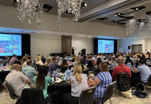 Participants at a large Hopefull Handbags Global team building event, listening to a speaker as they prepare to pack handbags with essentials for domestic abuse survivors.