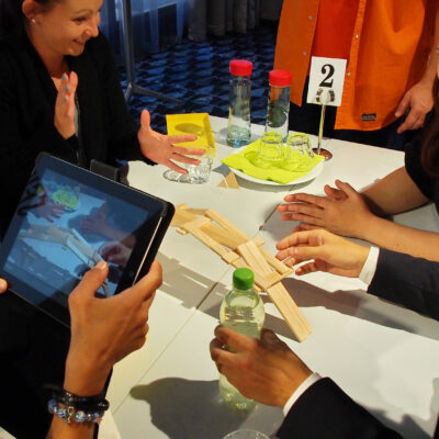 A team gathers around an iPad while engaging in a hands-on challenge, working together to complete a task in the Quickfire team building event. thumbnail