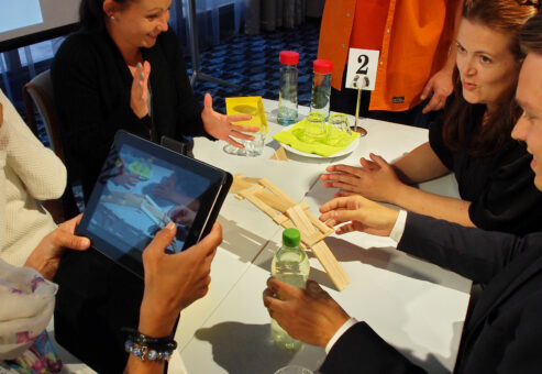 A team gathers around an iPad while engaging in a hands-on challenge, working together to complete a task in the Quickfire team building event.