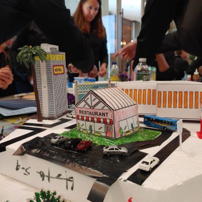Close-up of a miniature city model featuring a restaurant, hotel, parking lot, and roadway, created during a City Build team building activity. thumbnail