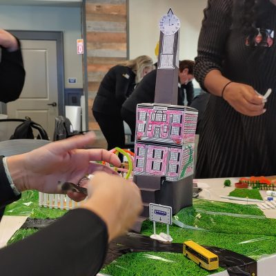 Close-up of a creative tower structure with a clock and decorated buildings in a City Build team building event, surrounded by miniature roads and greenery. thumbnail