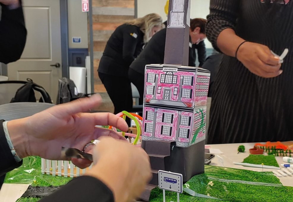 Close-up of a creative tower structure with a clock and decorated buildings in a City Build team building event, surrounded by miniature roads and greenery.