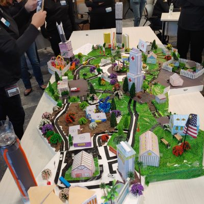 Participants at a City Build team building event admire and photograph an intricately designed miniature cityscape featuring roads, buildings, trees, and a central clock tower. thumbnail