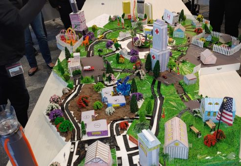 Participants at a City Build team building event admire and photograph an intricately designed miniature cityscape featuring roads, buildings, trees, and a central clock tower.