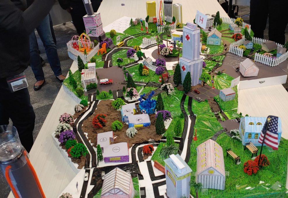 Participants at a City Build team building event admire and photograph an intricately designed miniature cityscape featuring roads, buildings, trees, and a central clock tower.