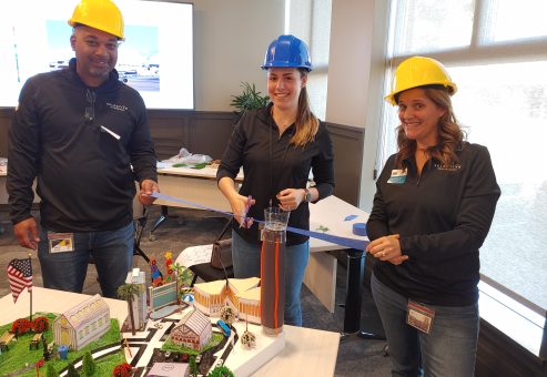Three team members in hard hats proudly cutting a ribbon to unveil their collaborative City Build project, featuring a detailed miniature urban landscape.