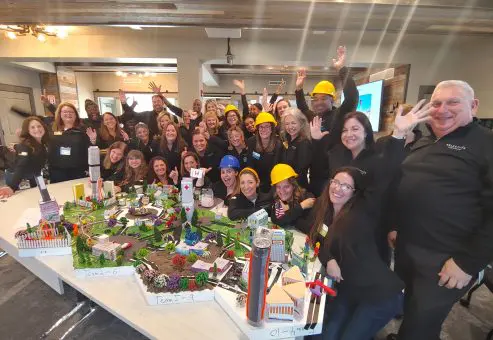 A large group of participants celebrates their success at a City Build team building event, posing around a detailed miniature city model while wearing hard hats and smiling enthusiastically.