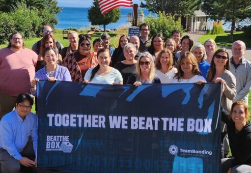 Group of colleagues proudly pose with a 'Together We Beat the Box' banner, celebrating their teamwork and success in the Beat the Box team building challenge.