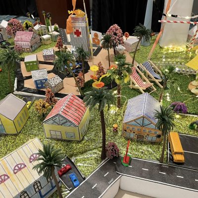 Close-up view of a vibrant City Build team building project showcasing miniature houses, trees, a school bus, and decorative lights on a grassy layout. thumbnail