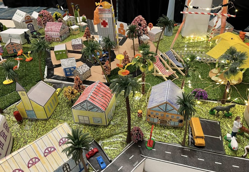 Close-up view of a vibrant City Build team building project showcasing miniature houses, trees, a school bus, and decorative lights on a grassy layout.