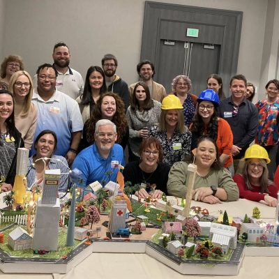Group photo of participants wearing hard hats, proudly posing behind their completed miniature city during a City Build team building event. thumbnail